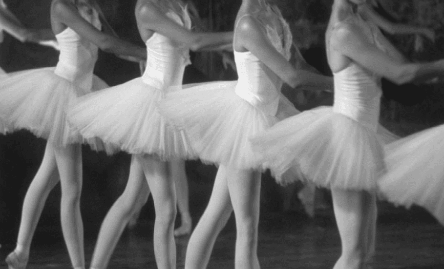 A line of ballerinas in white tutus performing synchronized dance steps in a black and white photograph
